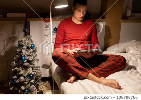 young man in christmas pyjama in bed watching a tablet young man in christmas pyjama in bed watching a tablet 106799766