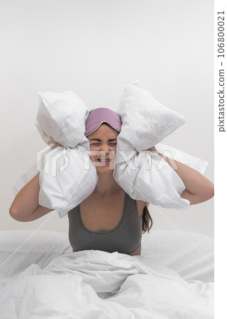 In distress, a woman covers her ears with pillows, unable to sleep due to disruptive neighbors, enduring a throbbing headache and insomnia. In distress, a woman covers her ears with pillows, unable to sleep due to disruptive neighbors, enduring a throbbing headache and insomnia. 106800021