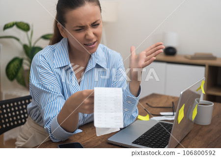 A surprised lady, homebound, confronting financial distress, holds a laptop, utility bill, and bank statements.  106800592