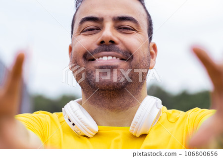 Smiling male with a bright t-shirt and wireless headphones has a great time, playfully framing the camera in a city setting taking selfie Smiling male with a bright t-shirt and wireless headphones has a great time, playfully framing the camera in a city setting taking selfie 106800656