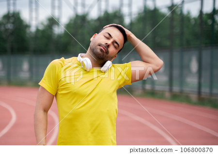 Man in yellow t-shirt doing neck side bend preparing muscles for athletic training on running track at city sports ground basic physical exercise for warming up 106800870