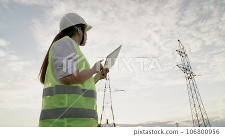 Woman engineer with helmet inputs data on tablet standing by electric power transmission line. Electrician with mobile device inspects power lines in field. Engineer at electric power substation Woman engineer with helmet inputs data on tablet standing by electric power transmission line. Electrician with mobile device inspects power lines in field. Engineer at electric power substation 106800956