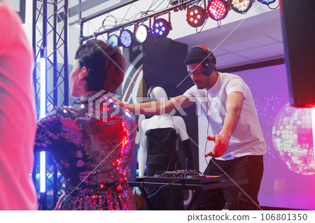 Young man dj mixing techno tracks on stage while people partying on dancefloor in nightclub. Musician playing electronic music on stage while clubbers dancing at discotheque Young man dj mixing techno tracks on stage while people partying on dancefloor in nightclub. Musician playing electronic music on stage while clubbers dancing at discotheque 106801350