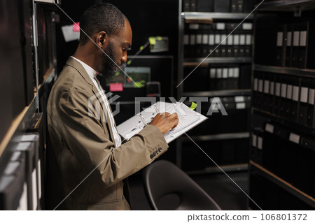 Investigator studying folder with police records in archive in office. African american detective standing near shelf, working late and analyzing forensic reports at night time 106801372