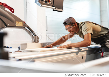 Young carpenter cutting a piece of wood in using a circular saw in furniture factory 106803246