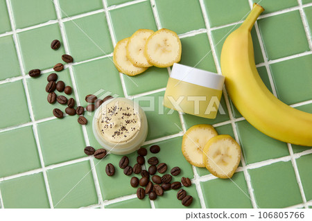 A yellow cream jar decorated with fresh banana, banana slices and brown coffee beans on green ceramic background. Mockup scene for cosmetic skin care, face mask and hair mask 106805766