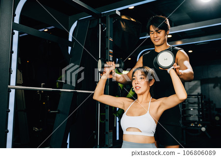 Asian fitness instructor teaching a smiling woman proper lifting techniques, using weight lifting equipment for a safe and effective workout, technique of exercise in gym dark gym background Asian fitness instructor teaching a smiling woman proper lifting techniques, using weight lifting equipment for a safe and effective workout, technique of exercise in gym dark gym background 106806089