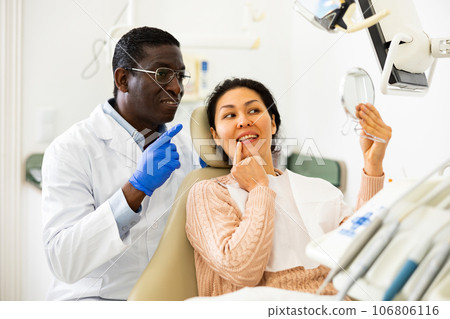 Smiling woman sitting and looking at mirror after tooth restoration in clinic 106806116
