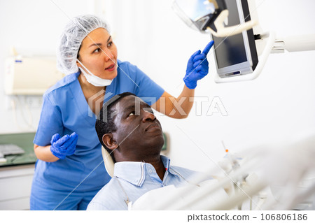 Man sitting on dental chair, female dentist pointing at display Man sitting on dental chair, female dentist pointing at display 106806186