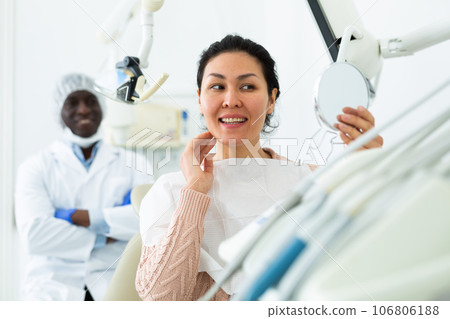 Woman satisfied with result of teeth treatment looking in mirror at dental office Woman satisfied with result of teeth treatment looking in mirror at dental office 106806188