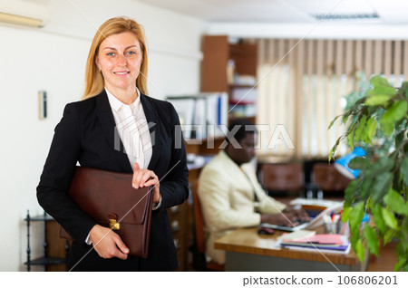 Smiling business woman with briefcase of documents standing in office 106806201