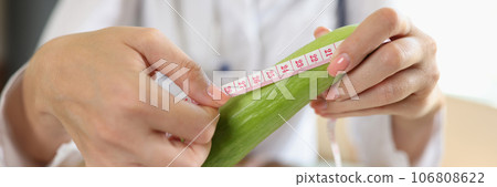 Doctor measures length of cucumber with measuring tape while sitting at table in medical clinic. Doctor measures length of cucumber with measuring tape while sitting at table in medical clinic. 106808622