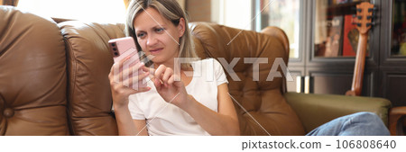 Concentrated woman looking at screen of her phone while sitting on couch at home. 106808640