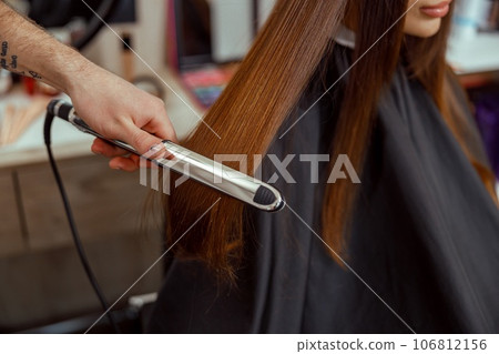 Closeup of hairdresser hand holding hair straightener and making hairstyle for female client 106812156