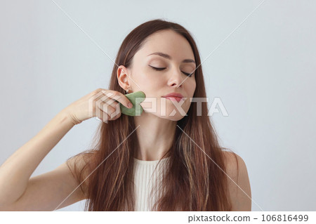 Close-up portrait of a beautiful girl, red-haired woman massaging her face with a stone scraper. Youth Corner 106816499