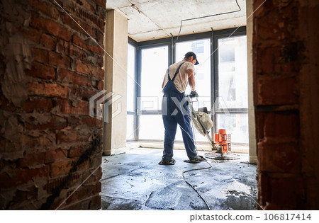Man finishing concrete surface with floor screed grinder machine in room with large window. Back view of male worker using troweling machine while screeding floor in apartment under renovation. 106817414