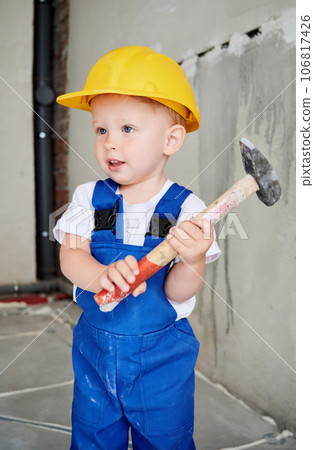 Adorable child construction worker holding hammer building repair tool. Cute kid wearing safety helmet and work overalls while posing at home during renovation. Adorable child construction worker holding hammer building repair tool. Cute kid wearing safety helmet and work overalls while posing at home during renovation. 106817426