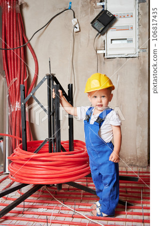 Full length of adorable little boy standing near underfloor heating pipes and looking at camera. Cute kid plumber wearing work overalls and safety helmet while posing in apartment under renovation. Full length of adorable little boy standing near underfloor heating pipes and looking at camera. Cute kid plumber wearing work overalls and safety helmet while posing in apartment under renovation. 106817435