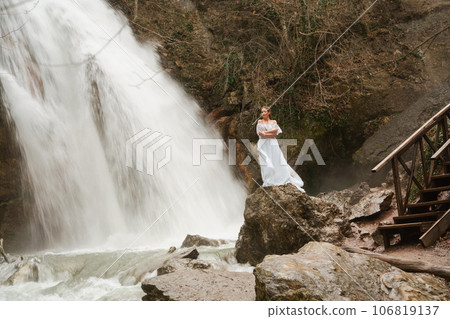 Happy woman in a white dress stands on a stone with a waterfall behind 106819137