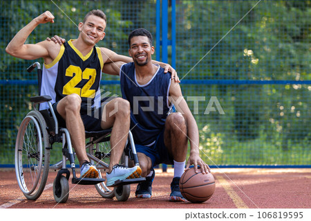 Man in wheelchair playing basketball with friend at outdoor court. 106819595