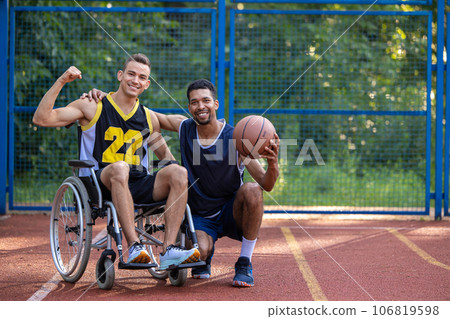 Man playing basketball with disabled friend in wheelchair at outdoor court. 106819598