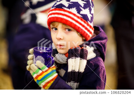 Little cute kid boy with cup of steaming hot chocolate or children punch. Happy child on Christmas market in Germany. Preschool kid in winter clothes warming hands on cup and drinking eggnog. 106820204