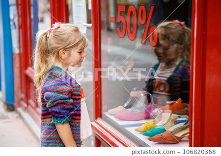 Little Girl looks at the window near the shopping center. Adorable happy child looking on shoes in store window. 106820368