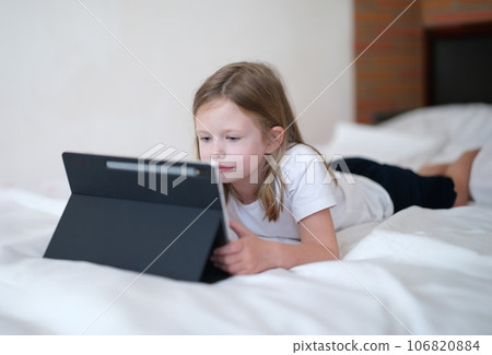 Little girl carefully looks at tablet while lying on bed 106820884