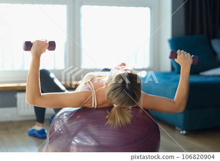 Young woman doing bench press with dumbbells and stabilizing ball at home 106820904