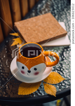 Autumn cozy still life on balcony, terrace. Fall cozy set with autumn fall leaves, cup of coffee or tea on brown books and notepads on balcony, terrace glass table. Relaxing still life concept. Autumn cozy still life on balcony, terrace. Fall cozy set with autumn fall leaves, cup of coffee or tea on brown books and notepads on balcony, terrace glass table. Relaxing still life concept. 106823012