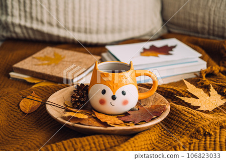 Autumn cozy home. Fall cozy reading nook with a blanket, candles, autumn fall leaves, cup of coffee on brown bed at home background. Cold season morning coffee, relaxing still life concept. 106823033
