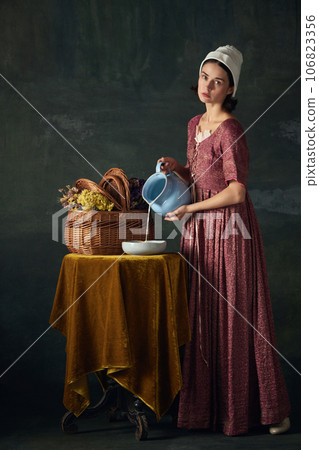 Countryside morning. Beautiful woman, medieval maid in historical attire pouring milk against vintage green background 106823356