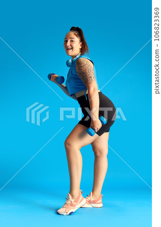 Young woman with overweight in sportswear training, doing exercises with dumbbells against blue background. Weight loss Young woman with overweight in sportswear training, doing exercises with dumbbells against blue background. Weight loss 106823369