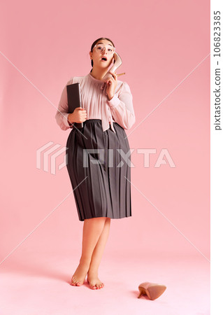 Woman in blouse and skirt standing barefoot, using heeled shoes like a mobile phone against pink background 106823385