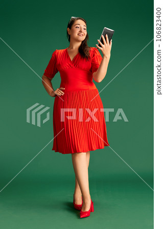 Full-length of beautiful woman in red dress looking on tablet, reading messages against green studio background 106823400