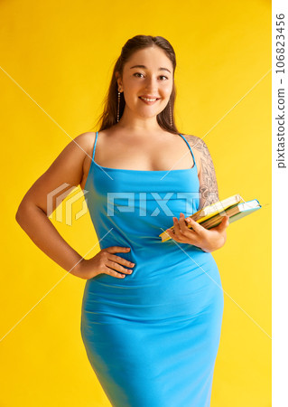 Portrait beautiful woman in blue dress posing books against bright yellow studio background. Smiling, delightful 106823456