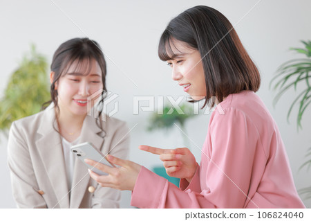 Two young women chatting while looking at their smartphones_1 Two young women chatting while looking at their smartphones_1 106824040