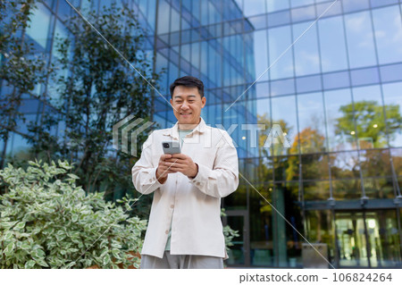 Successful Asian man in casual clothes uses the phone, man walks in the city near a modern building, dials a message, calls, businessman after work. Successful Asian man in casual clothes uses the phone, man walks in the city near a modern building, dials a message, calls, businessman after work. 106824264