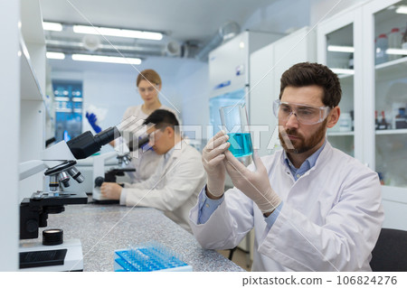 A young male scientist and chemist sits in a laboratory at a table and concentrates on studying a blue substance in a flask. Behind, male and female colleagues are working under a microscope. 106824276