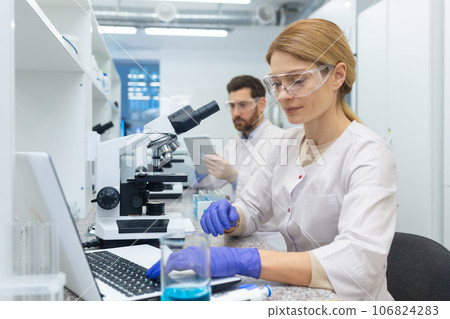 A young woman works in a laboratory, sits at a table with equipment and works on a laptop. Behind, a male colleague is using a tablet. 106824283