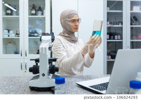 A Muslim young woman chemist and scientist in a hijab sits in a laboratory in front of a microscope and studies a blue liquid in a flask that she holds in her hands. 106824293