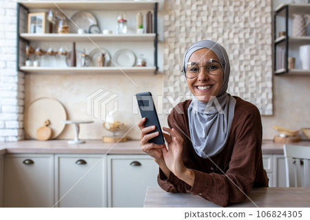 Portrait of young muslim woman in hijab smiling looking at camera, standing in kitchen at home and using mobile phone. Portrait of young muslim woman in hijab smiling looking at camera, standing in kitchen at home and using mobile phone. 106824305