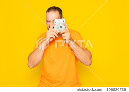Bearded Hispanic man in his 40s wearing an orange t-shirt taking a photograph with an instant camera, isolated on yellow studio background. Bearded Hispanic man in his 40s wearing an orange t-shirt taking a photograph with an instant camera, isolated on yellow studio background. 106826786