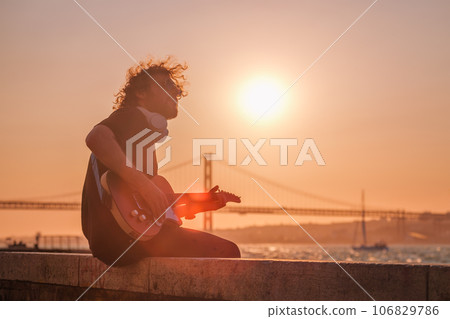 Hipster street musician in black playing electric guitar in the street on sunset on embankment with 25th of April bridge in background. Lisbon, Portugal 106829786