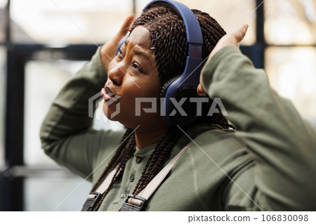 Happy supervisor wearing headset listening music during work break in warehouse, dancing and having fun. African american employee with industrial overall doing goods quality control in storehouse 106830098