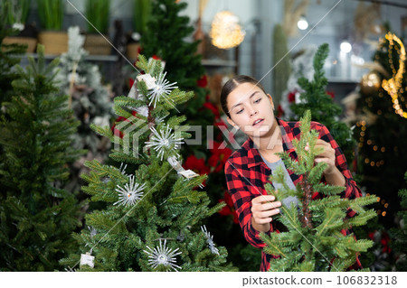 Young female in plaid shirt buying artificial Christmas trees before holidays in a store 106832318