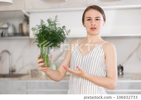 Woman in dress with short sleeves sitting near kitchen table and holding glass with bunch of dill Woman in dress with short sleeves sitting near kitchen table and holding glass with bunch of dill 106832733