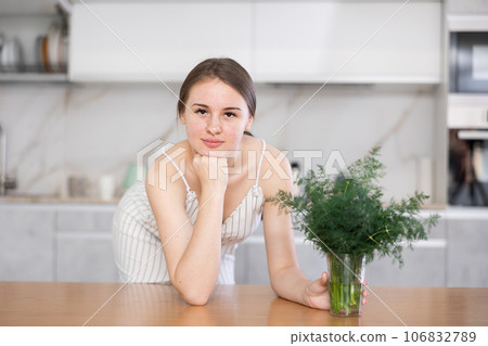 Woman in dress with short sleeves sitting near kitchen table and holding glass with bunch of dill Woman in dress with short sleeves sitting near kitchen table and holding glass with bunch of dill 106832789
