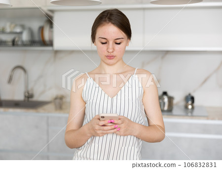 Young girl standing in the kitchen and looking at her phone Young girl standing in the kitchen and looking at her phone 106832831