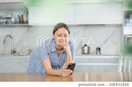 In the kitchen, teen woman leans on table with phone in hand, propping up chin with fist. 106832888